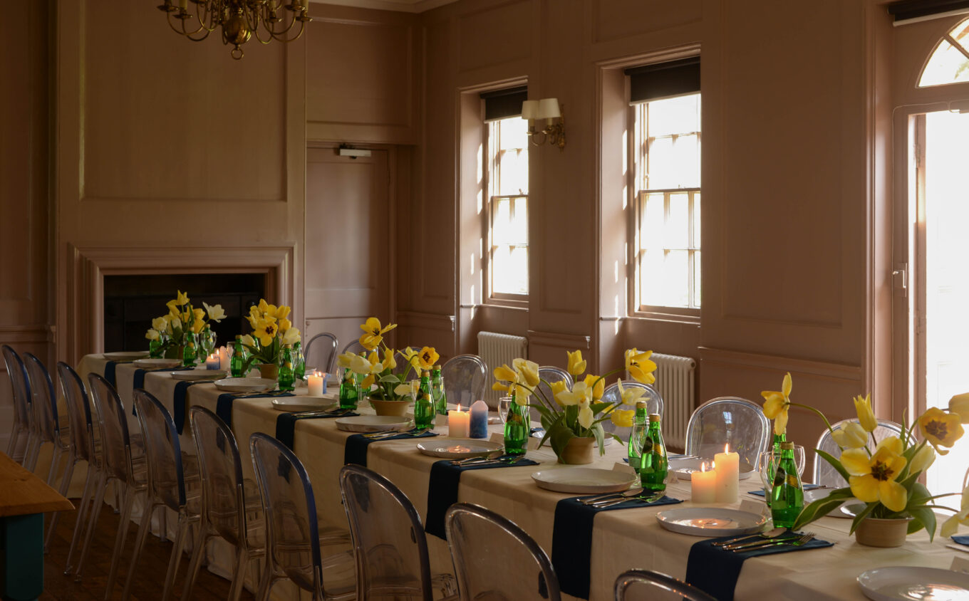 A Georgian period room with large windows and sun streaming onto a long table dressed for dinner with daffodils decorating it