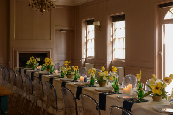 A Georgian period room with large windows and sun streaming onto a long table dressed for dinner with daffodils decorating it