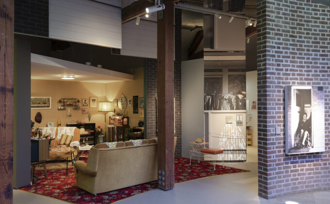 A distant installation view showing two adjacent recreated domestic scenes under an industrial gallery roof. On the left is A Terraced House in 1978, with mustard yellow wallpaper, red patterened carpet, and various decors on the wall and cabinet. On the right is a glimpse of the entry to A High-rise Flat in 2005 with a white wall with multiple doorbells. A large black-and-white portrait of a person in a wheelchair is mounted on the adjacent brick wall.
