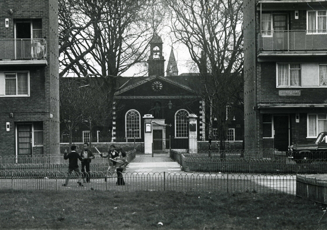 A black and white urban scene showing brick apartment buildings flanking the Geffrye's Museum's clock tower. Children play in the fenced courtyard area between the buildings, with bare winter trees framing the central structure.