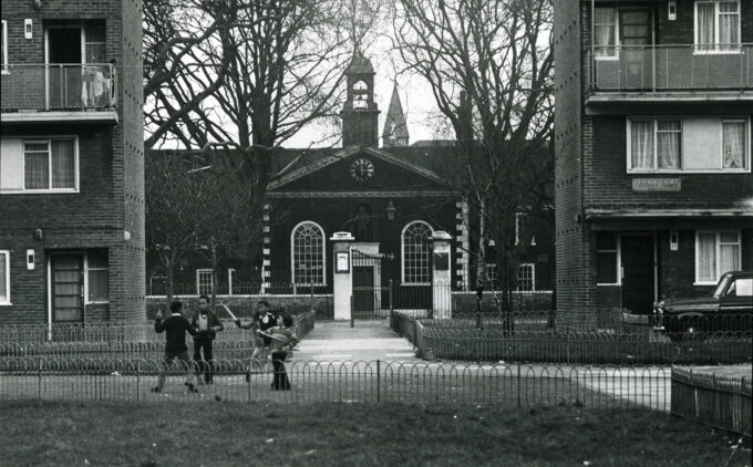 A black and white urban scene showing brick apartment buildings flanking the Geffrye's Museum's clock tower. Children play in the fenced courtyard area between the buildings, with bare winter trees framing the central structure.