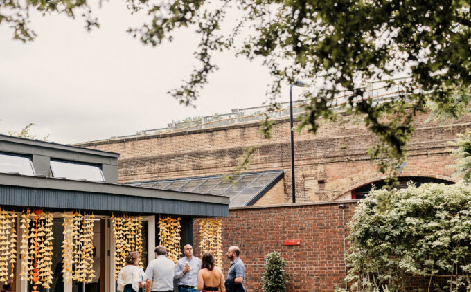 "A small group of people gathering outside a modern building with large windows and decorative lighting, set against a brick wall backdrop with a landscaped garden featuring potted plants and herbs in the foreground.