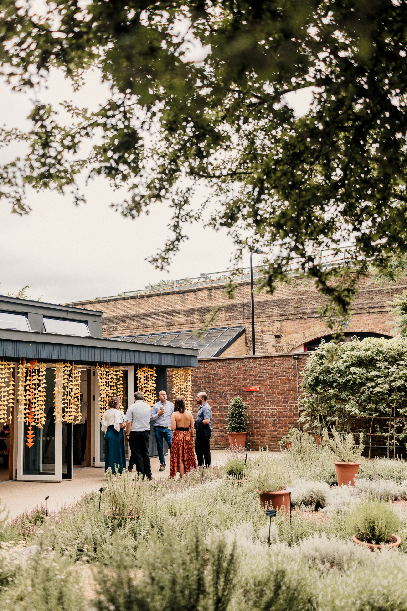 "A small group of people gathering outside a modern building with large windows and decorative lighting, set against a brick wall backdrop with a landscaped garden featuring potted plants and herbs in the foreground.