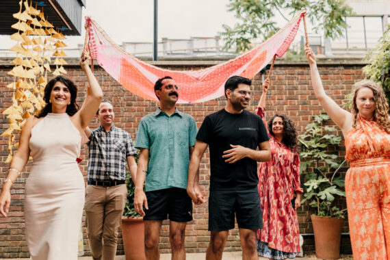 A marriage ceremony in a brick-walled courtyard: the couple stands centre front holding hands in joy, while four people at the corners around them smile and hold a pink patterned cloth overhead. Behind them are hanging yellow decorations.