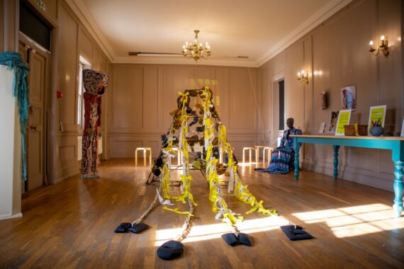 An airy gallery with sunlight on wood floors. A central rope-and-yellow-ribbon sculpture trails to weighted anchors, surrounded by low stools, textiles, and fabric-clad mannequin figures by a turquoise table.