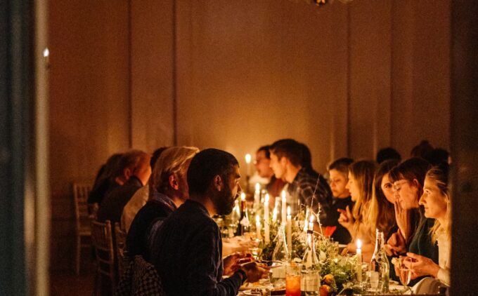 A candlelit supper with guests eating, chatting, and smiling as they sit along a long banquet table set with sparkling glassware, flickering tapers, and seasonal greenery under a chandelier.