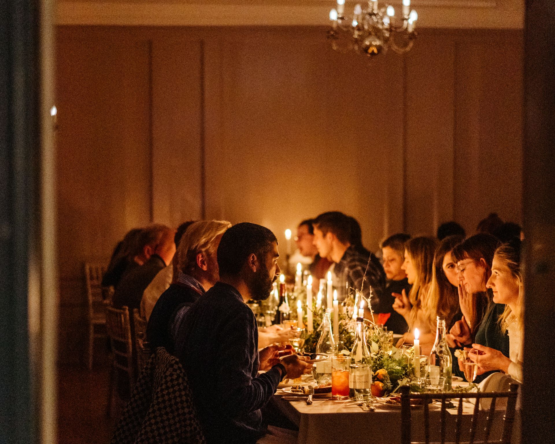 A candlelit supper with guests eating, chatting, and smiling as they sit along a long banquet table set with sparkling glassware, flickering tapers, and seasonal greenery under a chandelier.