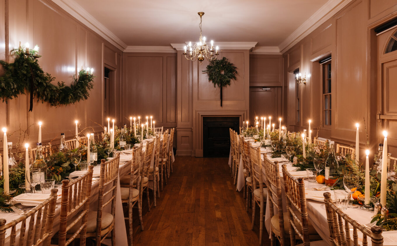 Long dining tables set for a supper with lit taper candles, greenery, and oranges. The wood-panelled room is decorated with festive wreaths and warm lighting from chandeliers and wall sconces.