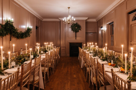 Long dining tables set for a supper with lit taper candles, greenery, and oranges. The wood-panelled room is decorated with festive wreaths and warm lighting from chandeliers and wall sconces.