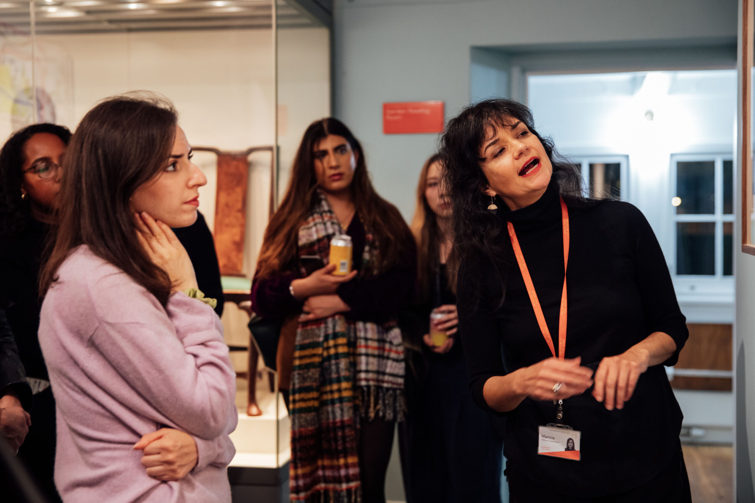A Visitor Experience host is leading a tour at a evening event in a gallery, with young adults listening intently.