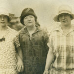 Sepia-toned photograph of three women standing arm-in-arm. Each wears a cloche hat and a patterned top - the central figure, the mother, in a darker hat and geometric print, two daughters in lighter hats and floral and plaid dresses respectively.