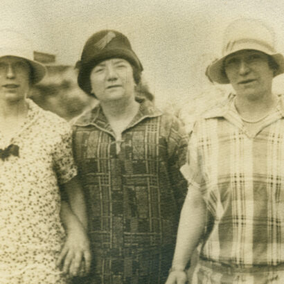 Sepia-toned photograph of three women standing arm-in-arm. Each wears a cloche hat and a patterned top - the central figure, the mother, in a darker hat and geometric print, two daughters in lighter hats and floral and plaid dresses respectively.