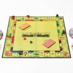 A square lime yellow Monopoly board laid flat on a white background. Two dice (one red, one blue) rest in the centre. On both sides of the board are stacks cards and paper money in various denominations. Various colours of tokens sit on different areas on the board.