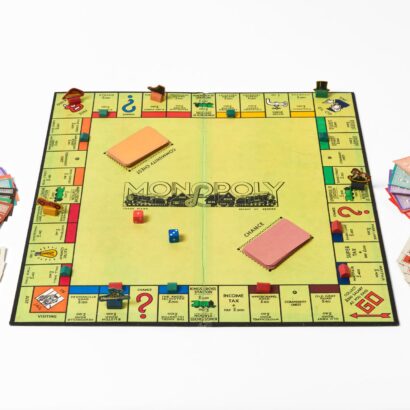 A square lime yellow Monopoly board laid flat on a white background. Two dice (one red, one blue) rest in the centre. On both sides of the board are stacks cards and paper money in various denominations. Various colours of tokens sit on different areas on the board.