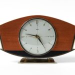 Mechanical mantel clock on a white background: reddish-brown horizontal trapezoid case on two small brass feet above a small dark-brown plinth, central circular white dial with gold 12-3-6-9 numerals and dot hour markers, hands at 9:25:30.