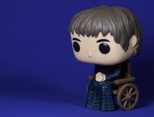 A Funko Pop! figure of Bran Stark from Game of Thrones, seated in a brown wooden wheelchair, with a large head and minimalist facial features, on a deep blue backdrop