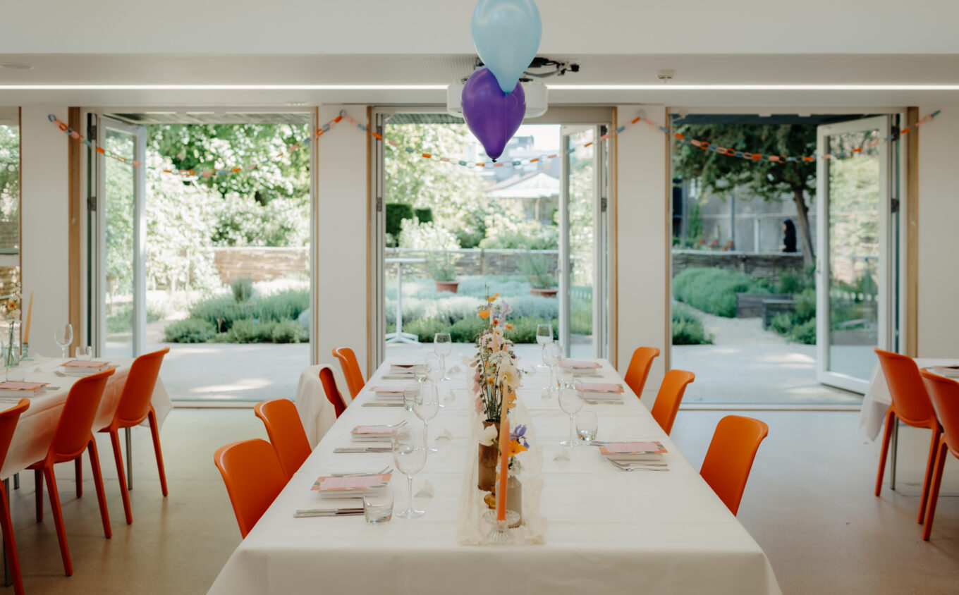 A table in a large room laid for a party with orange seats and a blue and purple balloon
