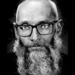 Black and white photo of a man with a beard and glasses staring at the camera.