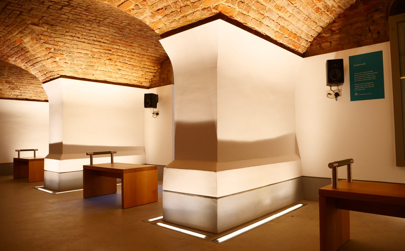 A dimly lit room with grey floor, white walls and a low, curved brick celling. Black speakers are visible.