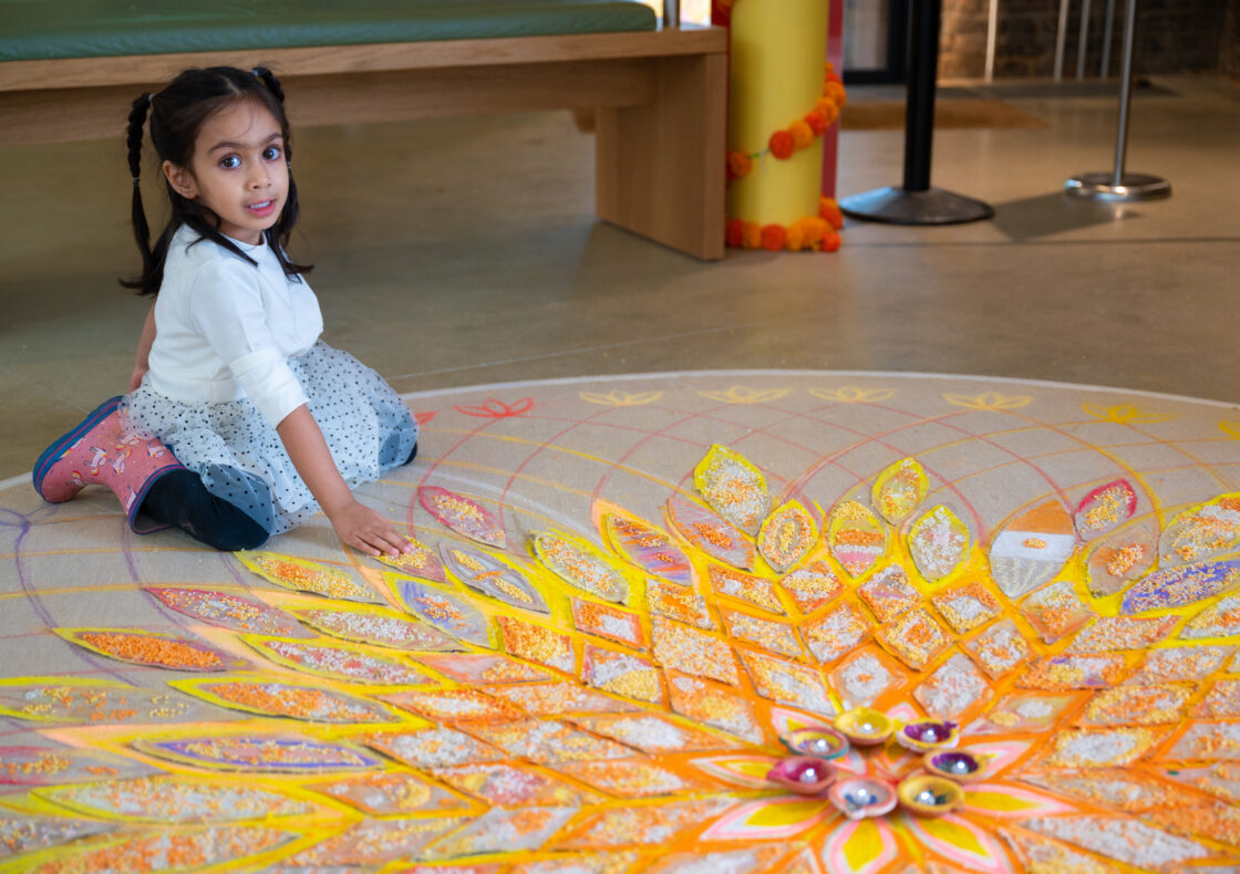 A child kneels beside a large Diwali rangoli drawing on the floor, her hand gently touching the edge of the pattern. The rangoli design is is drawn in orange, yellow, and purple chalk and filled with small beads. There is a wooden bench and a column wrapped with garlands in the background.