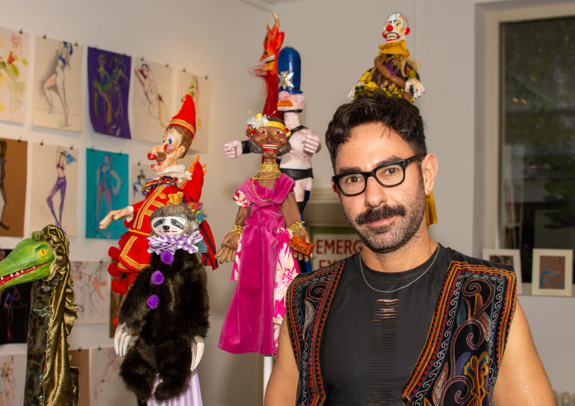 A man is wearing black-rimmed glasses and a patterned vest, standing in a bright gallery space. Surrounding him are several hand-crafted puppets on poles—a sloth in a purple ruff, a clown in a red costume, a figure in a pink dress, and a blue-helmeted character—against a backdrop of fashion sketches pinned to the wall.
