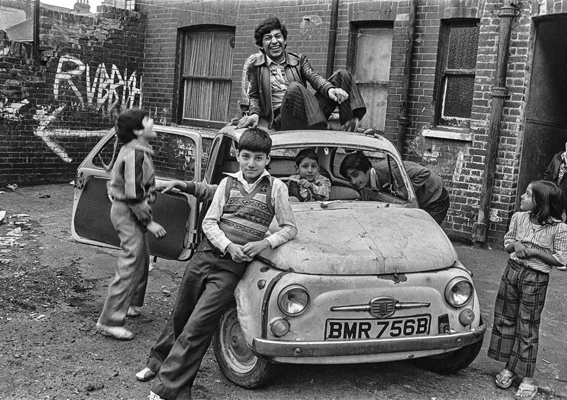A black-and-white photograph of children playing on and around an old Fiat in a brick-walled courtyard. Two peek out from the front window, while one sits atop the car laughing and another stands to the left, gazing up. A child leans against the bonnet with hands in his pockets, and another watches from the right as someone else laughs in the background. Graffiti scrawls adorn the wall behind them.