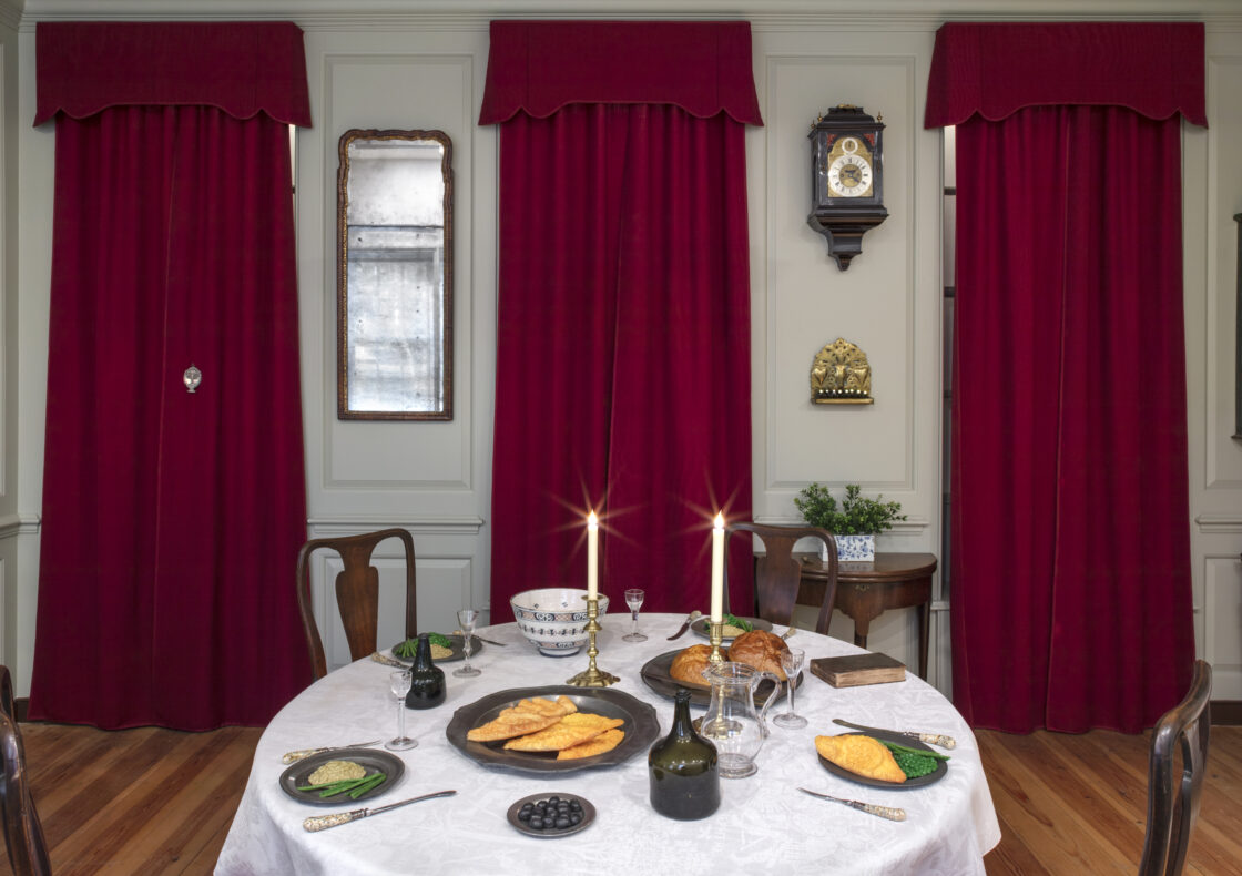 An installation view of a parlour in 1745 set for Hannukah at the Museum. There is a round table covered with a white fabric and set with two lit candlesticks, pastries and green beans, small glass goblets, and a prayer book. Behind it, three tall windows are dressed with deep red drapes. between the middle and right windows hang a clock. A narrow gilt mirror is mounted between the left and centre windows.