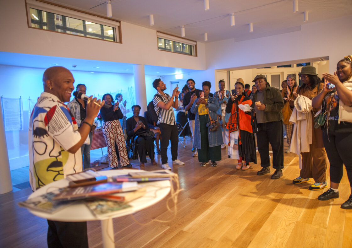 A man speaking into a microphone to a crowd of attendees listening in a modern white-walled space.