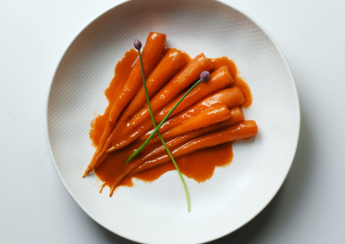 A white plate with slender glazed carrots in a vibrant orange sauce, garnished with two green chive stalks. Served on a minimalist white plate against a white background.