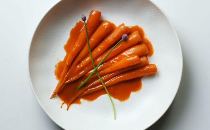 A white plate with slender glazed carrots in a vibrant orange sauce, garnished with two green chive stalks. Served on a minimalist white plate against a white background.