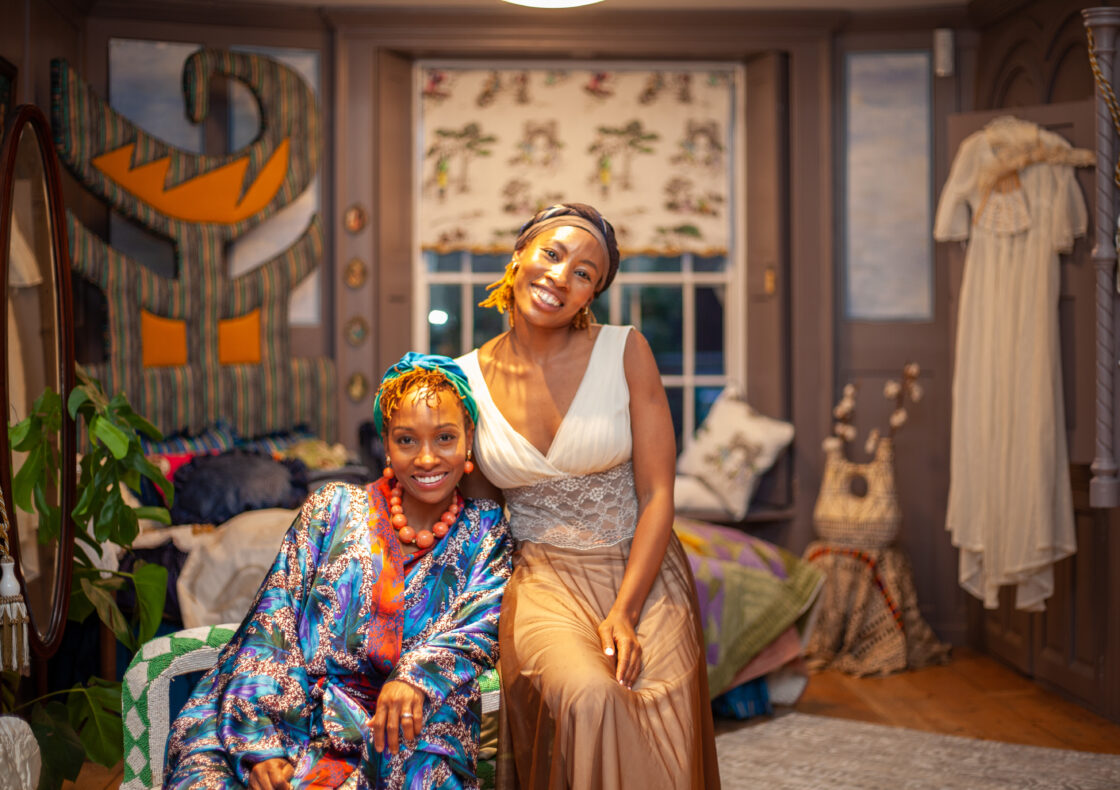 Two women pose and smile in a bright and decorative bedroom. One of them sits in a green and white armchair and the other sits besides her on one of the armrests. Behind them, the room is furnished with a bed draped in layered quilts, a full-length mirror, leafy houseplants, hanging garments, and patterned wall hangings
