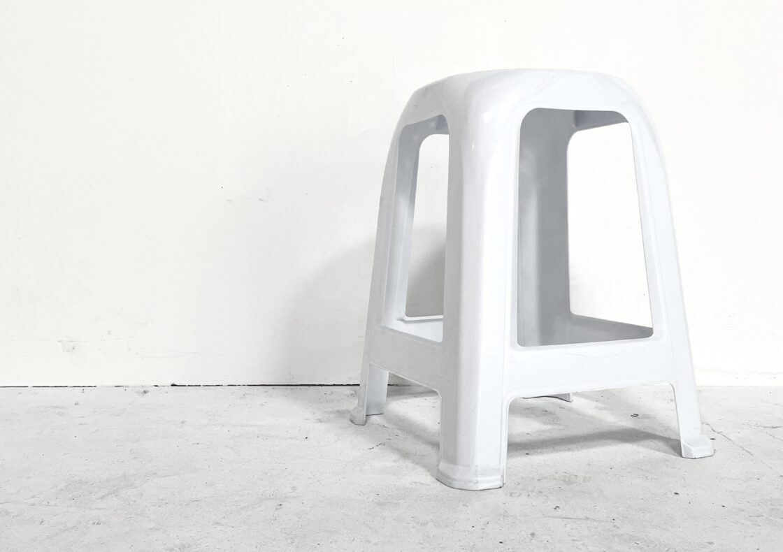 A white stool with four splayed legs stands alone on a pale concrete floor against a white wall.