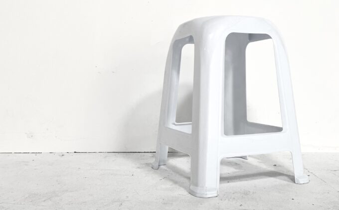 A white stool with four splayed legs stands alone on a pale concrete floor against a white wall.