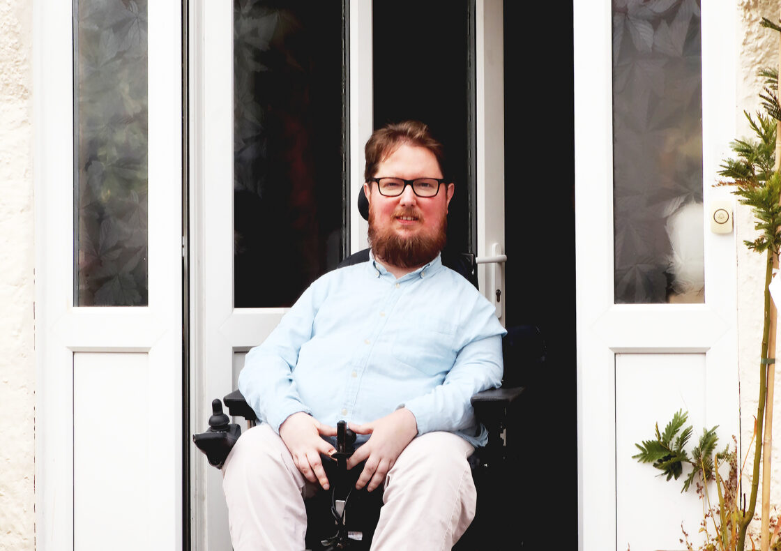 Person is sitting in a powered wheelchair in front of the doorway, with a gentle smile. They wear glasses, a light blue shirt, and beige trousers. The front door is slightly open, revealing a step free threshold with potted plants on either side.
