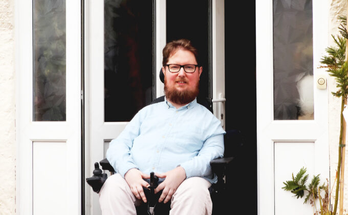 Person is sitting in a powered wheelchair in front of the doorway, with a gentle smile. They wear glasses, a light blue shirt, and beige trousers. The front door is slightly open, revealing a step free threshold with potted plants on either side.