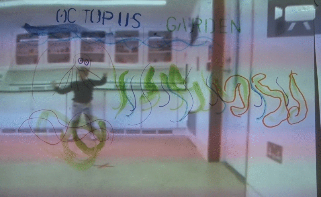 A room viewed through a translucent pane covered in child-made marker doodles and words. Across the top, “Octopus Garden” is written in blue and green. Below, looping wavy lines in green, blue, and orange swirl around a simple outline of a smiling octopus with waving tentacles. Beyond the pane, the blurred figure of a child stands in a white-walled gallery or classroom, hands raised, with cabinets lining the back wall.