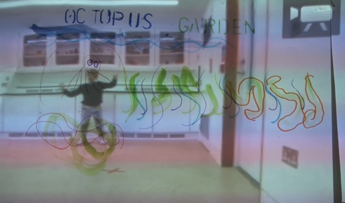 A room viewed through a translucent pane covered in child-made marker doodles and words. Across the top, “Octopus Garden” is written in blue and green. Below, looping wavy lines in green, blue, and orange swirl around a simple outline of a smiling octopus with waving tentacles. Beyond the pane, the blurred figure of a child stands in a white-walled gallery or classroom, hands raised, with cabinets lining the back wall.