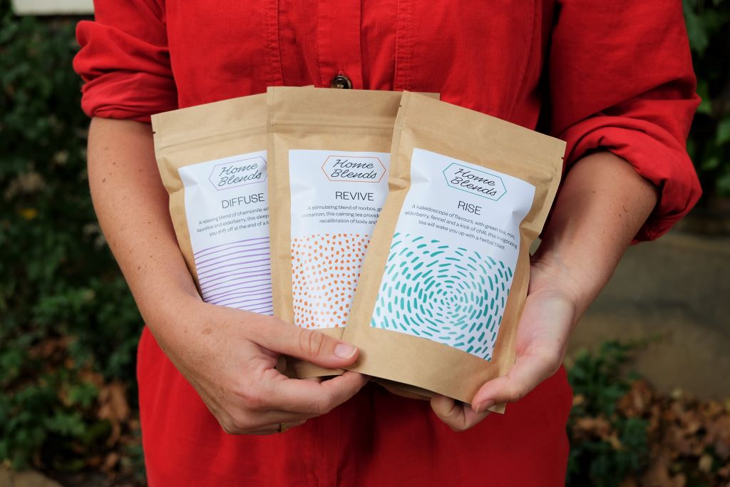 Hands holding three tea blend packages labeled 'DIFFUSE,' 'REVIVE,' and 'RISE' from Home Blends.