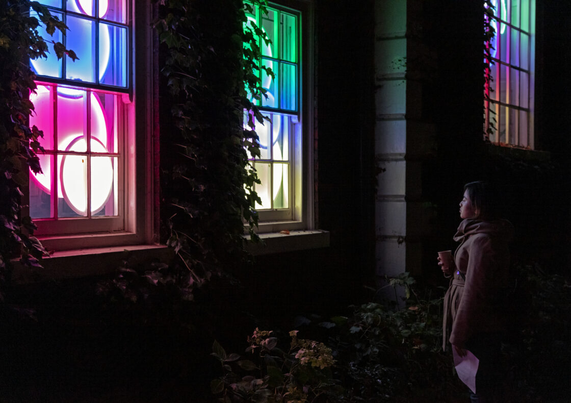 A woman stands outside at night, holding a takeaway cup and looking up at three tall windows framed by ivy. Behind the glass, an installation of overlapping neon circles glows in pink, blue, green, and yellow, casting coloured light onto the surrounding brick and foliage.