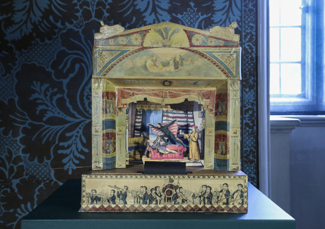 A fold out, three-dimensional paper toy theatre displayed on a green plinth. Its arch is richly painted with classical motifs in muted reds, blues, and golds. Inside, a miniature stage scene shows a winged imp hovering above a pale figure reclining on a pink chaise, while a robed character looks on. The front panel is illustrated with a line of musicians playing under a conductor