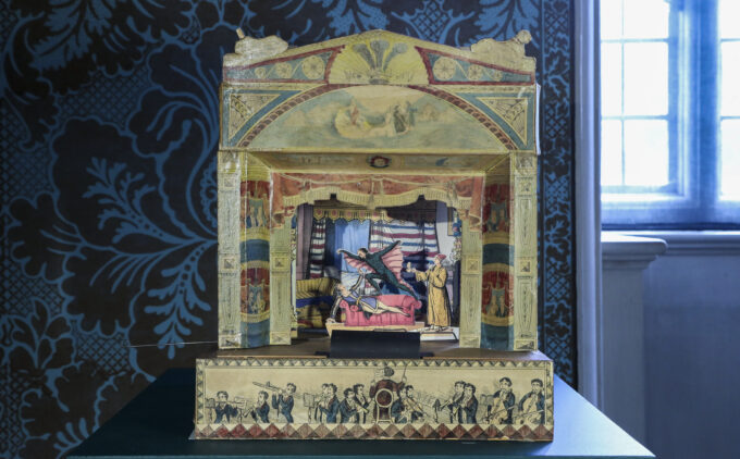 A fold out, three-dimensional paper toy theatre displayed on a green plinth. Its arch is richly painted with classical motifs in muted reds, blues, and golds. Inside, a miniature stage scene shows a winged imp hovering above a pale figure reclining on a pink chaise, while a robed character looks on. The front panel is illustrated with a line of musicians playing under a conductor