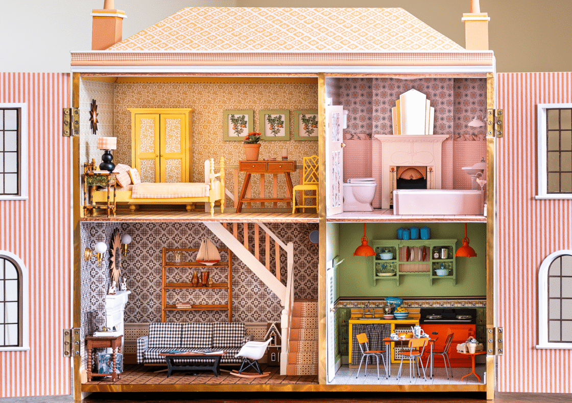 A fold out, two-story paper pink dollhouse displayed against a neutral background. The top left room is a yellow-toned bedroom with patterned wallpaper, a single bed, side table, and writing desk. To its right, a pink bathroom features a bathtub, toilet, sink, and decorative mantel. Below, the bottom left living room is clad in brown-and-white patterned wallpaper, with a sofa, rocking chair, fireplace, and shelving. The bottom right kitchen has green walls, orange AGA cooker and lighting, open shelving with dishes, and a small dining table with chairs.
