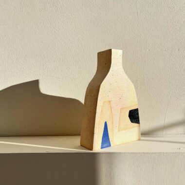 A geometric ceramic vase with a flat base and sloped shoulders, decorated with abstract forms in muted peach, blue, and black. Sunlight casts a strong shadow.