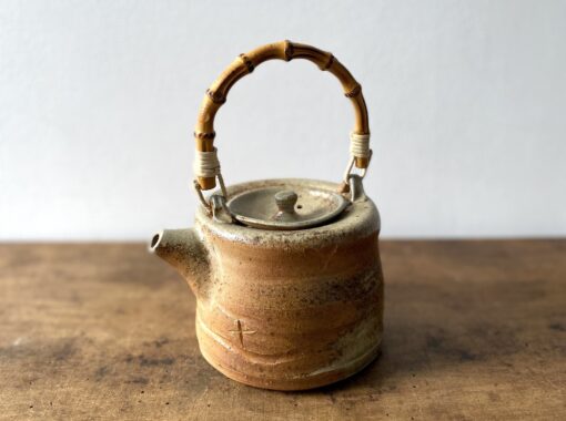 A rustic ceramic teapot with earthy tones and a round body sits on a wooden surface. It features a curved bamboo handle secured with white twine.