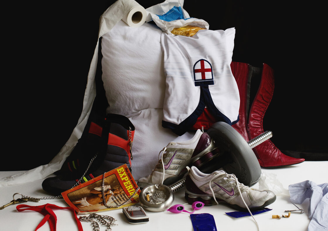 A still life of personal items including a pillow, red cowboy boots, sneakers, a dumbbell-part, red and black heeled boot, a pair of nike sneakers, toilet paper, Greggs wrapper, red thong with chains, jewelry, a condom packet, and a magazine, all stacked together on a white cloth against a black background.
