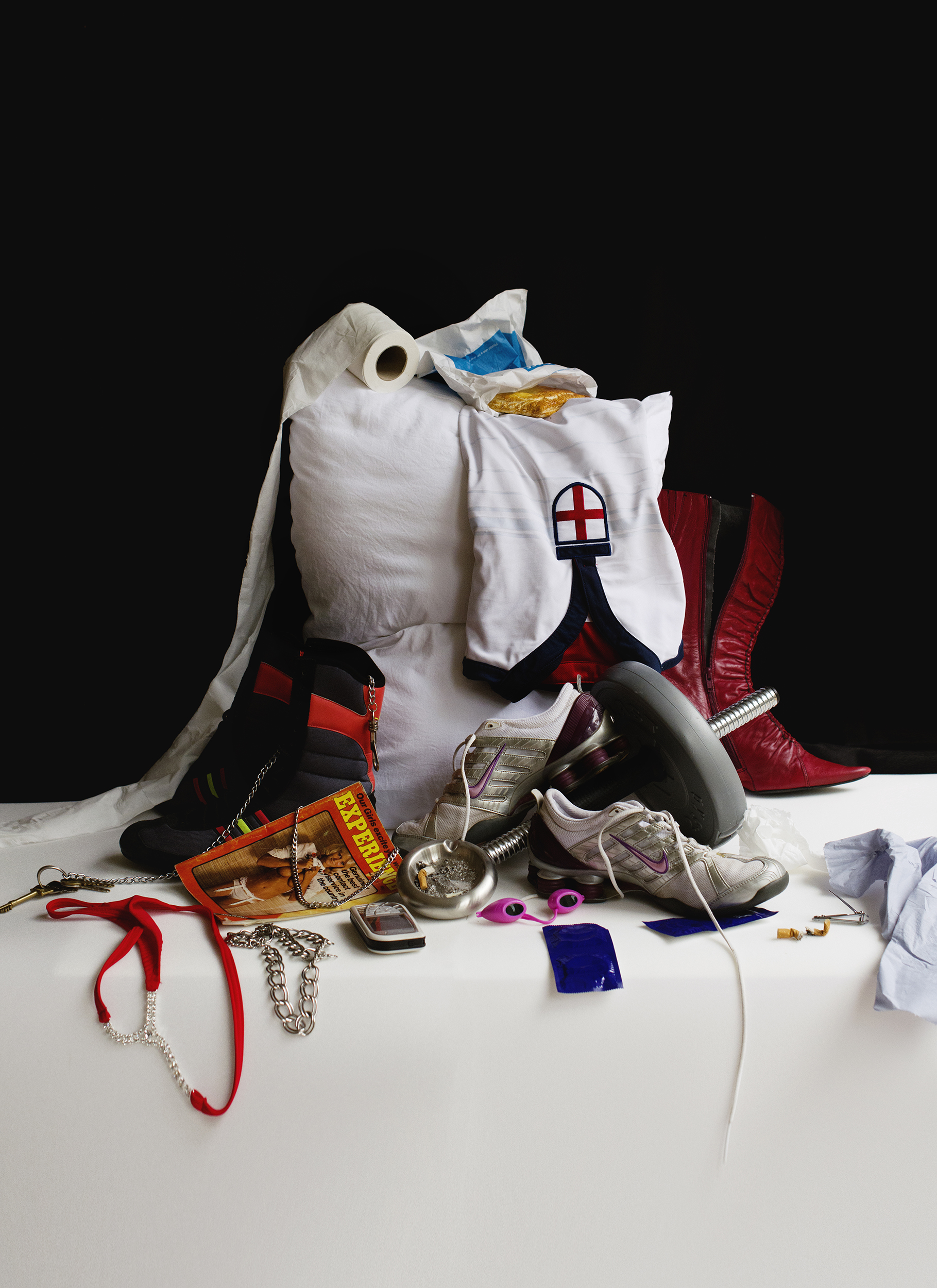 A still life of personal items including a pillow, red cowboy boots, sneakers, a dumbbell-part, red and black heeled boot, a pair of nike sneakers, toilet paper, Greggs wrapper, red thong with chains, jewelry, a condom packet, and a magazine, all stacked together on a white cloth against a black background.