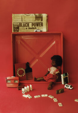 A red-toned still life featuring a vintage Black Power newspaper, a Black doll in a red dress, dominoes, toy vehicles, a pick comb, and a View-Master, all arranged in and around a red plastic tray.