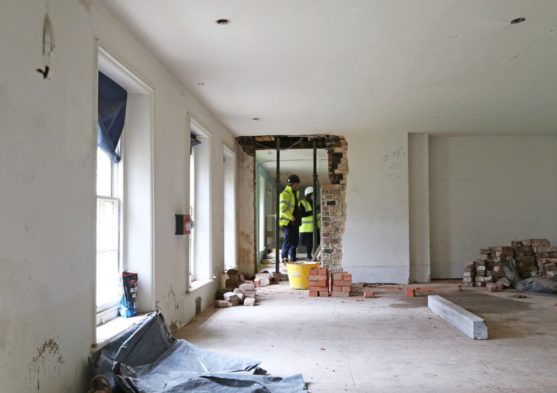 Inside a partially gutted room, construction workers in hi-vis jackets and helmets consult at an exposed brick wall opening. Piles of bricks, rubble, and windows line the space.