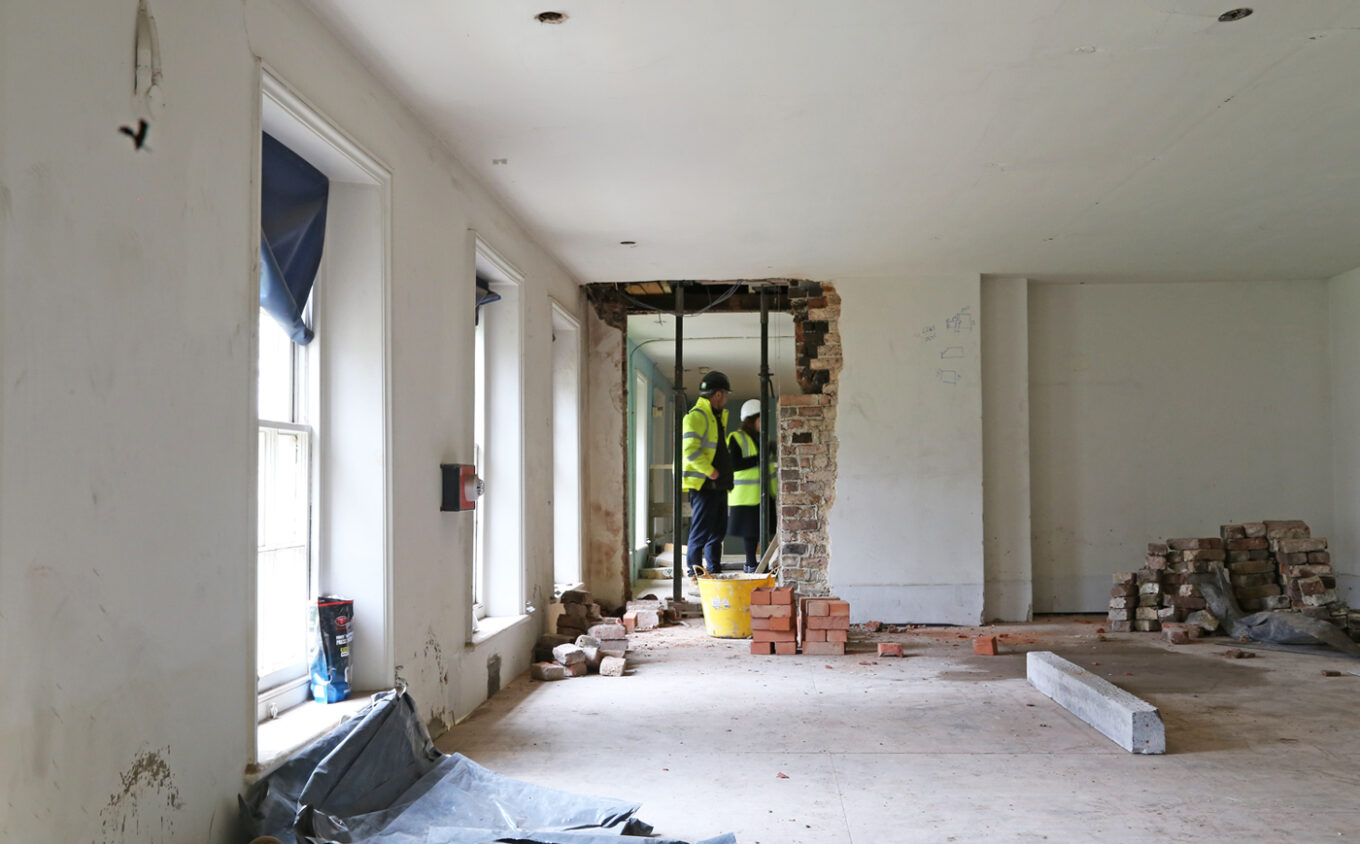 Inside a partially gutted room, construction workers in hi-vis jackets and helmets consult at an exposed brick wall opening. Piles of bricks, rubble, and windows line the space.