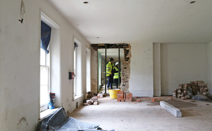 Inside a partially gutted room, construction workers in hi-vis jackets and helmets consult at an exposed brick wall opening. Piles of bricks, rubble, and windows line the space.
